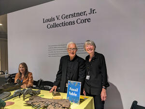 Two Museum volunteers pose behind a Fossil Table laid out with fossil specimens in front of wall with text "Louis V. Gerstner Jr. Collections Core."