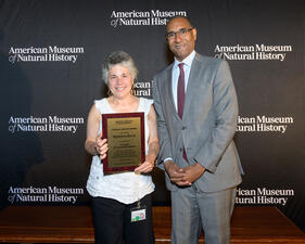 Museum volunteer Madeline Byrne stands beside Sean Decatur in front of a step and repeat holding a plaque honoring her service.