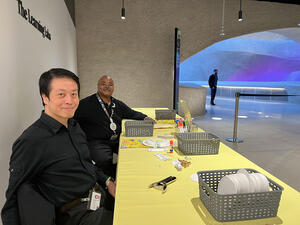 Museum volunteers Tung and Craig sit at a long table with arts and crafts materials on it in the Gilder Center.