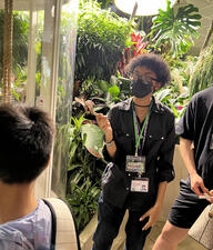 A Museum volunteer holds a cylindrical object with a butterfly perched on it and speaks to visitors in the Butterfly Vivarium.