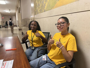 Two young volunteers in brightly colored AMNH shirts sit behind an information desk, both holding two thumbs up.