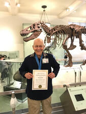 Dan Jacobs holds his certificate of appreciation in front of the T. rex mount in the Hall of Saurischian Dinosaurs.