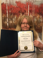 Eileen Flood stands in front of a forest painting, holding her certification of appreciation.