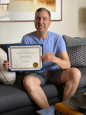James Berlanti sits on a couch holding his certificate of appreciation.
