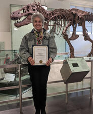 Myrella Triana stands in front of the T. rex skeleton, and holds a certificate honoring her 25 years of volunteer service.