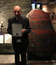 Paul Lewison stands in the Mignone Halls of Gems and Minerals, and holds a certificate honoring his 20 years of volunteer service.