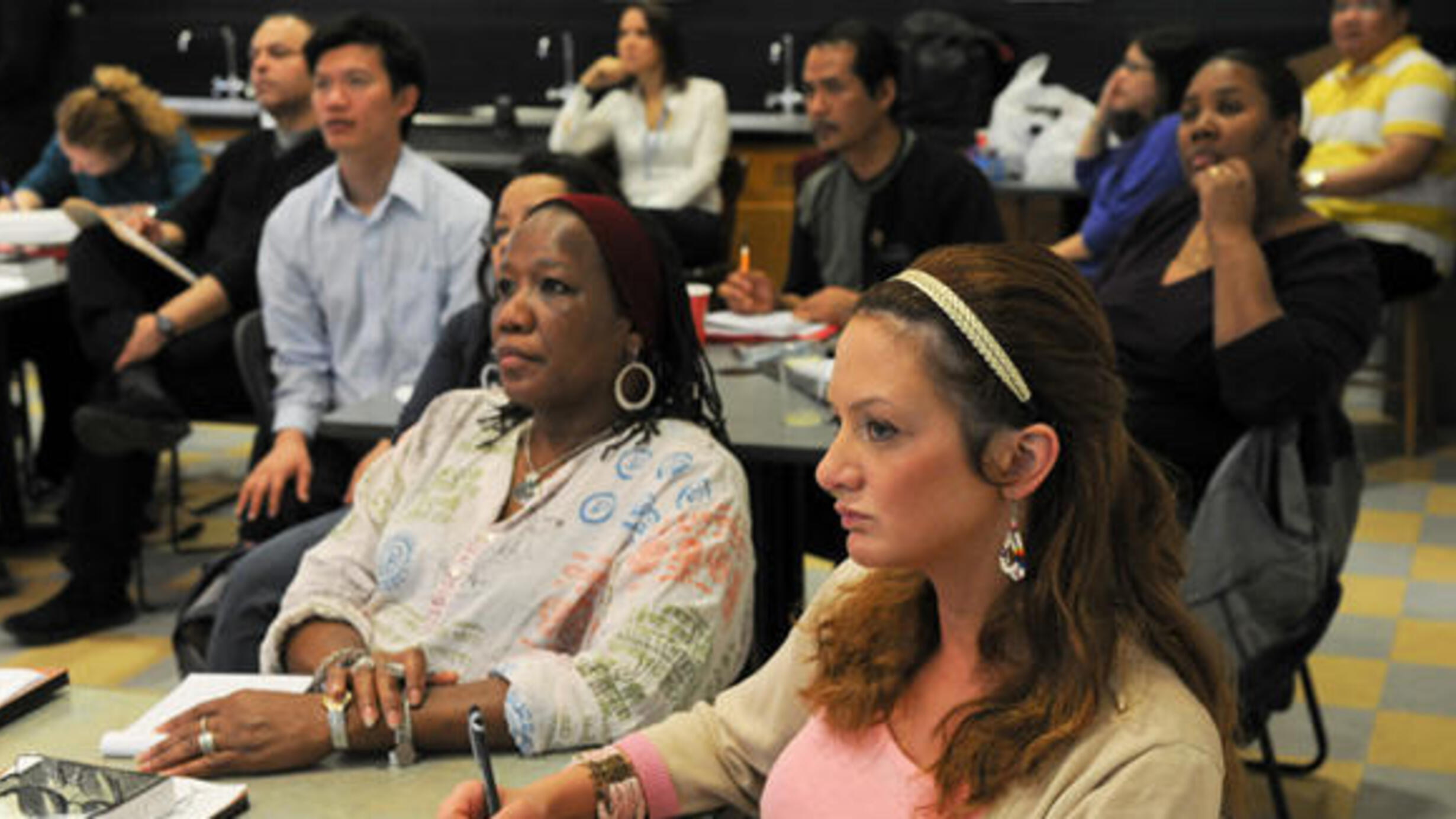 Ten people seated at four different tables, many with pens and notebooks in front of them, all look attentively toward the left.