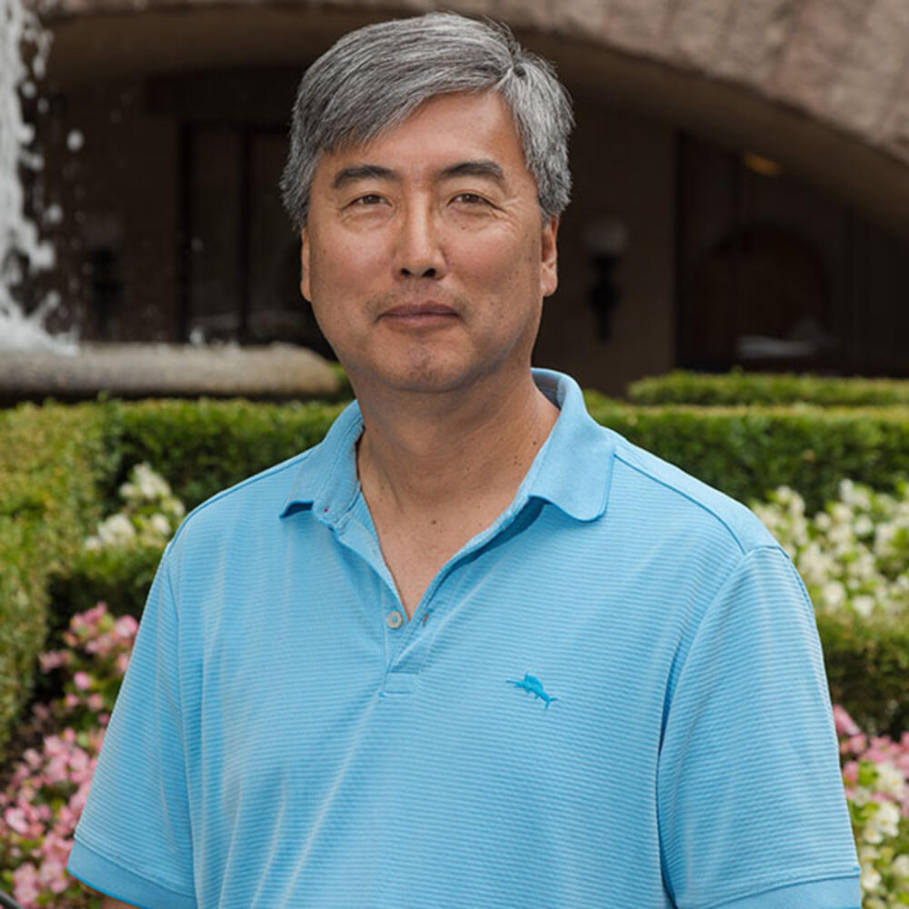 Namshik Yoo poses for a portrait outdoors, with a garden in view behind him.