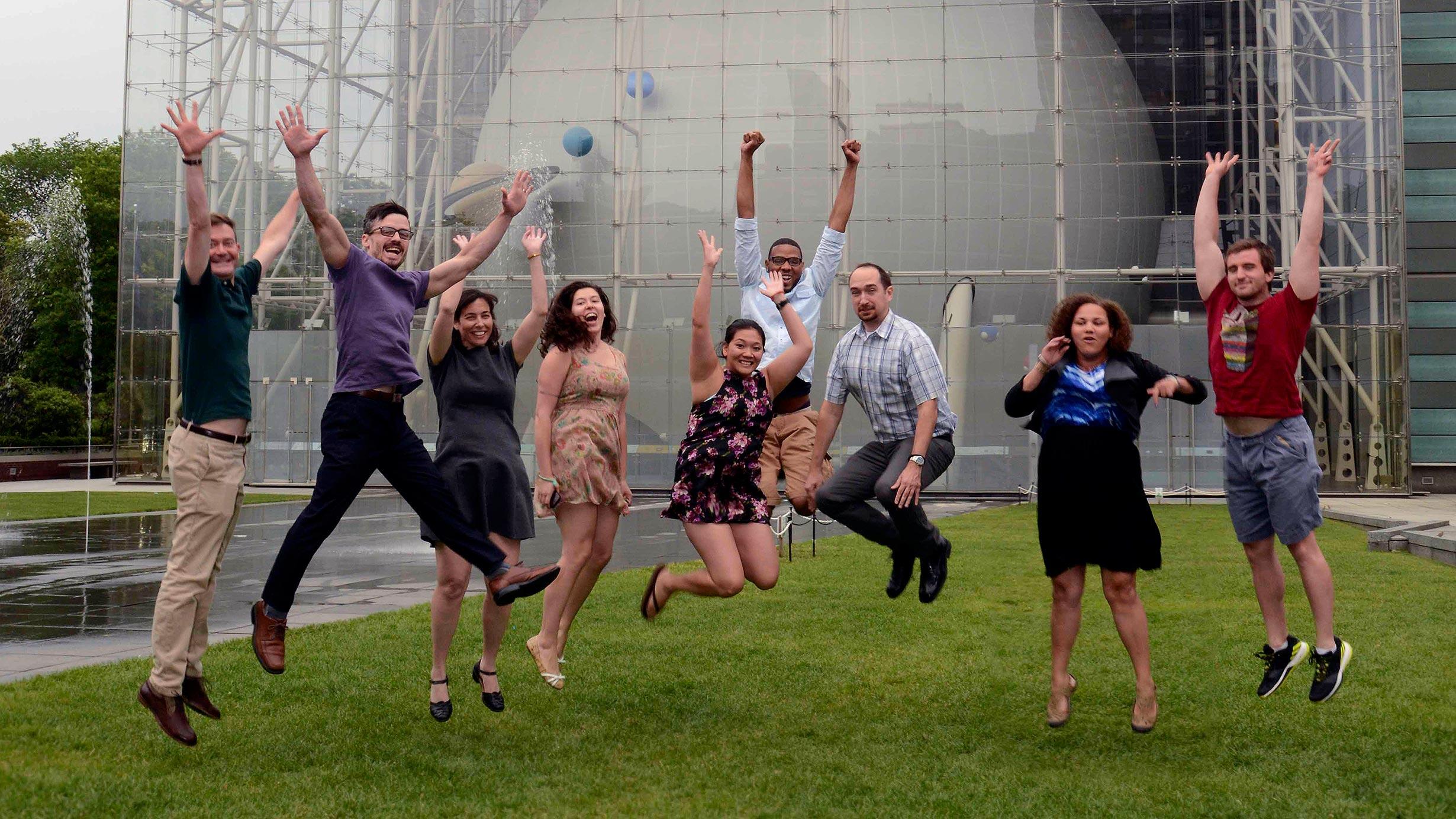 Nine students in the MAT Program jump in the air on the lawn in front of the Rose Center for Earth and Space.