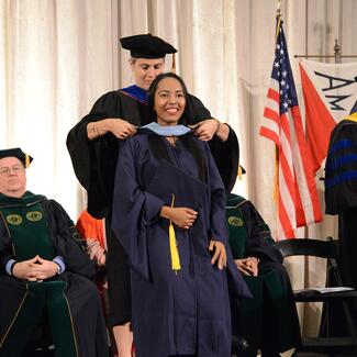 An MAT graduate is awarded a sash during the graduation ceremony, as other faculty members look on.