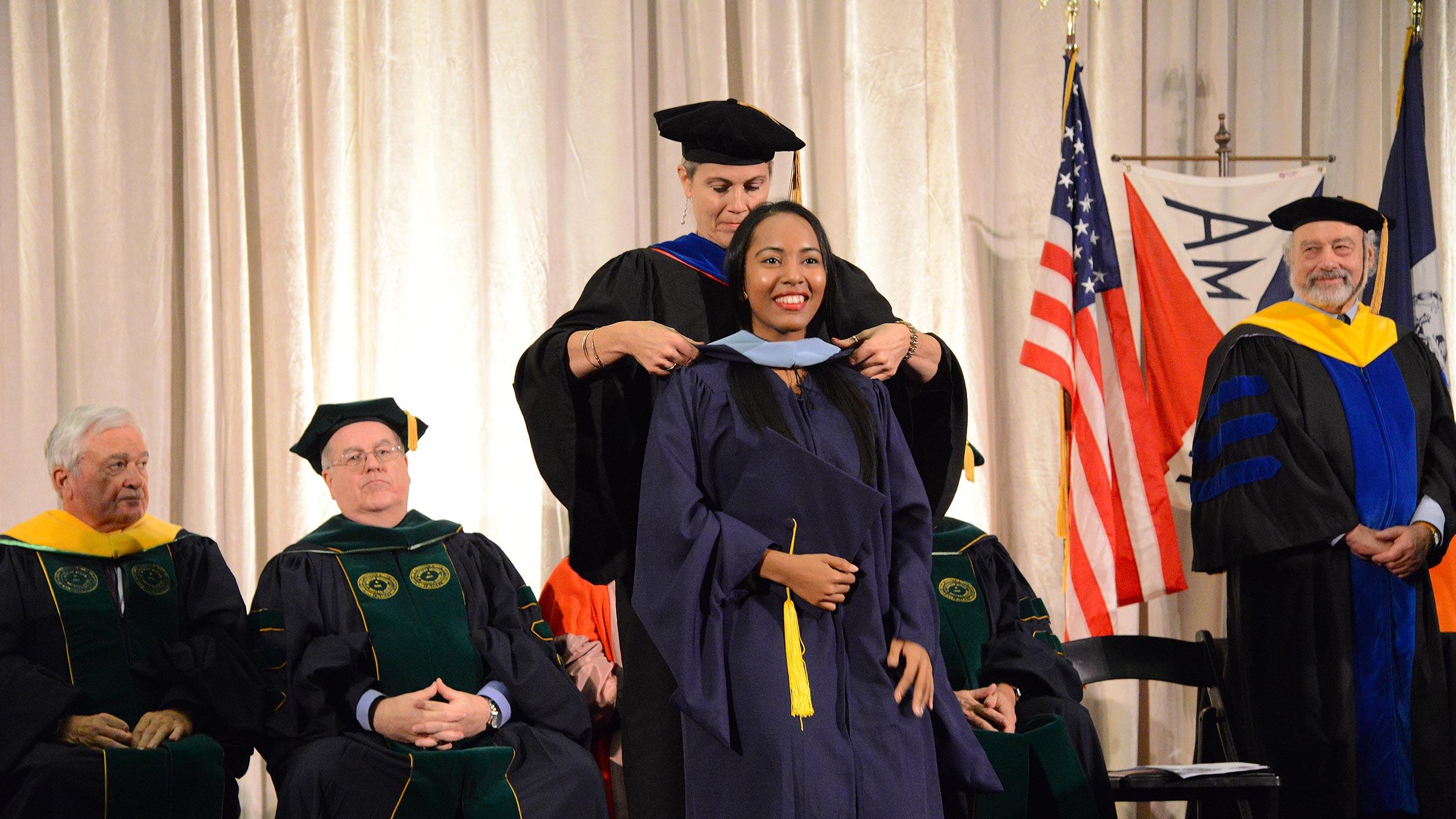 An MAT graduate is awarded a sash during the graduation ceremony, as other faculty members look on.