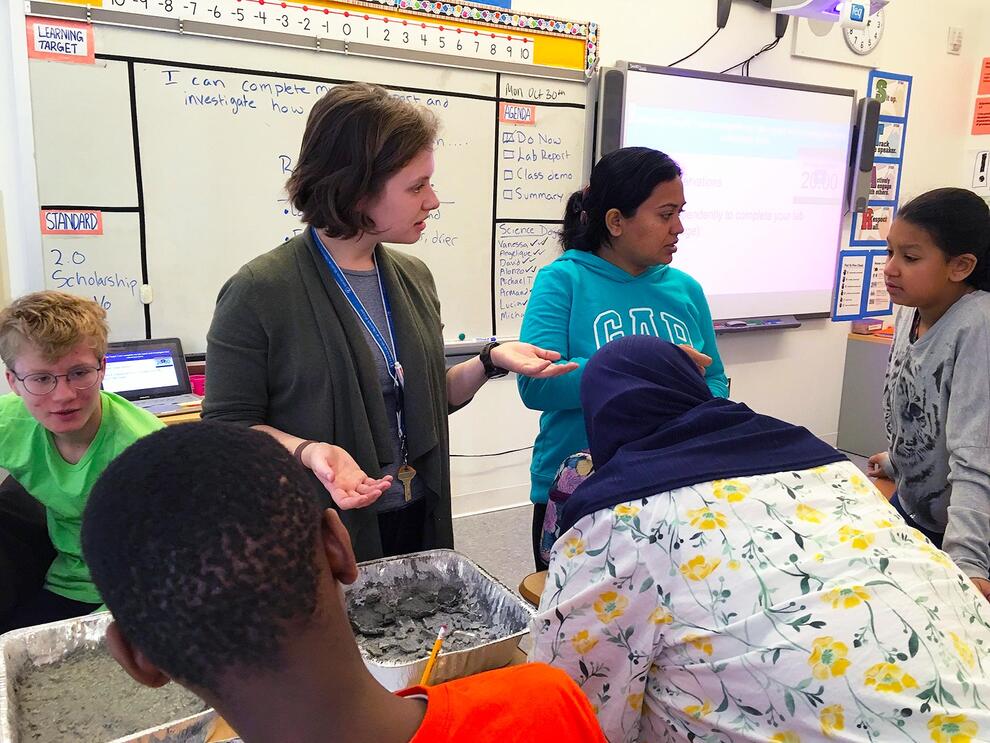 MAT Faculty member stands in a classroom and talks to several students.