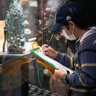 A child wearing a baseball cap and face mask stands in front of a rock specimen with a pencil and paper on a clipboard.
