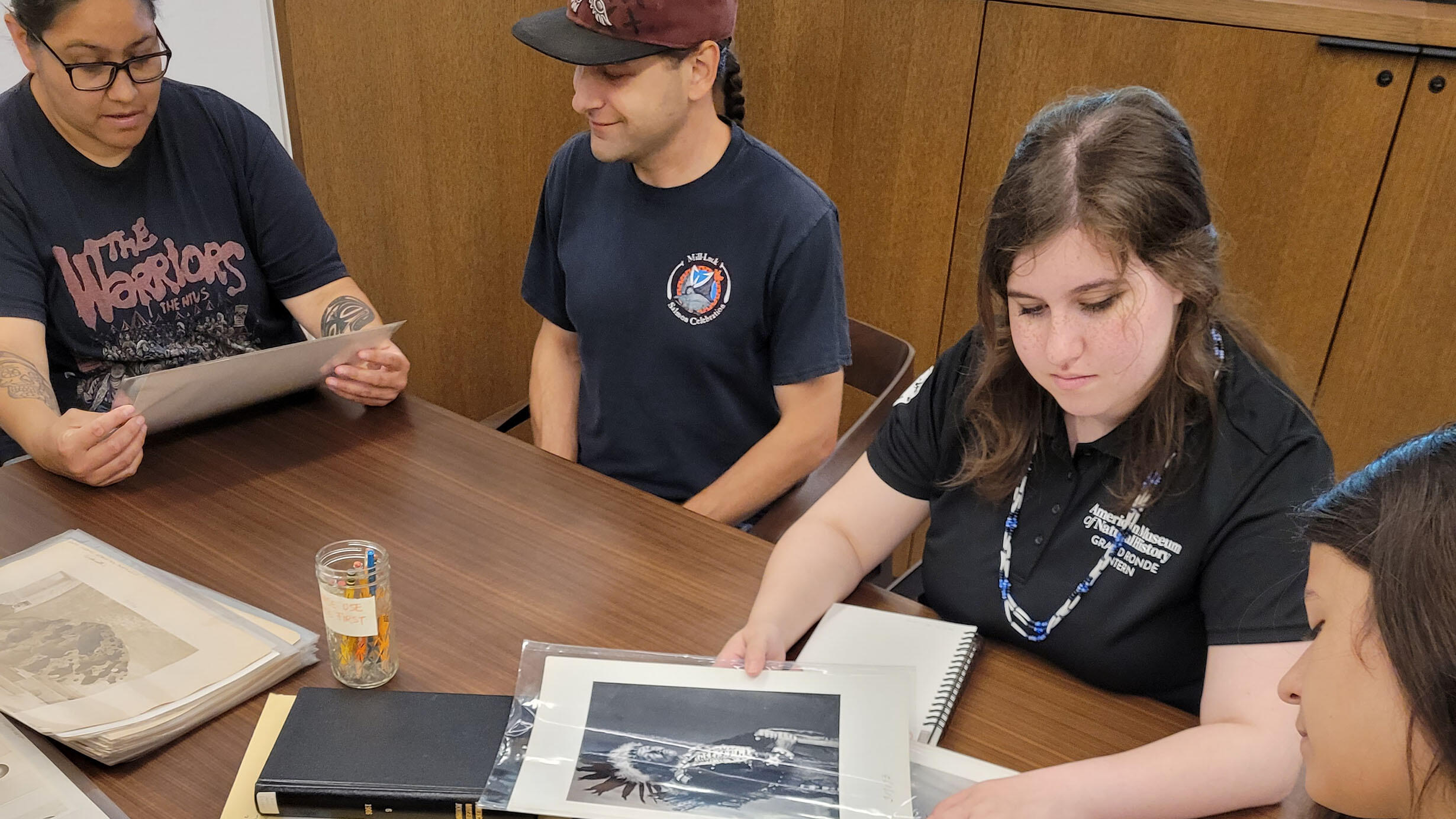 Four young people, including one wearing a polo embroidered with "AMNH - Grand Ronde Intern," sit around a table examining images in clear plastic folders.