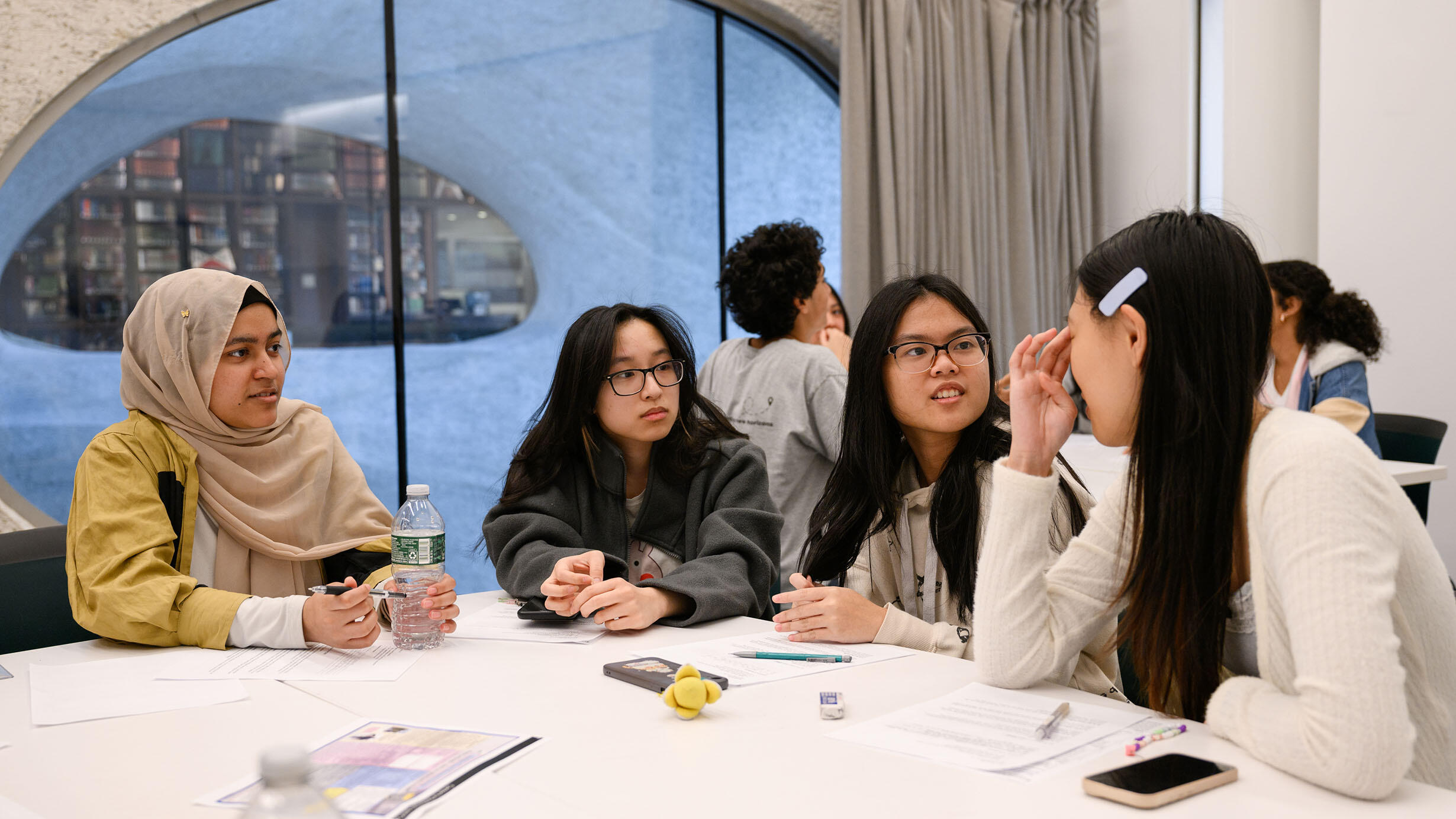 Four students sit at a table and talk inside a classroom, with a window view showing the Gottesman Library across the way.