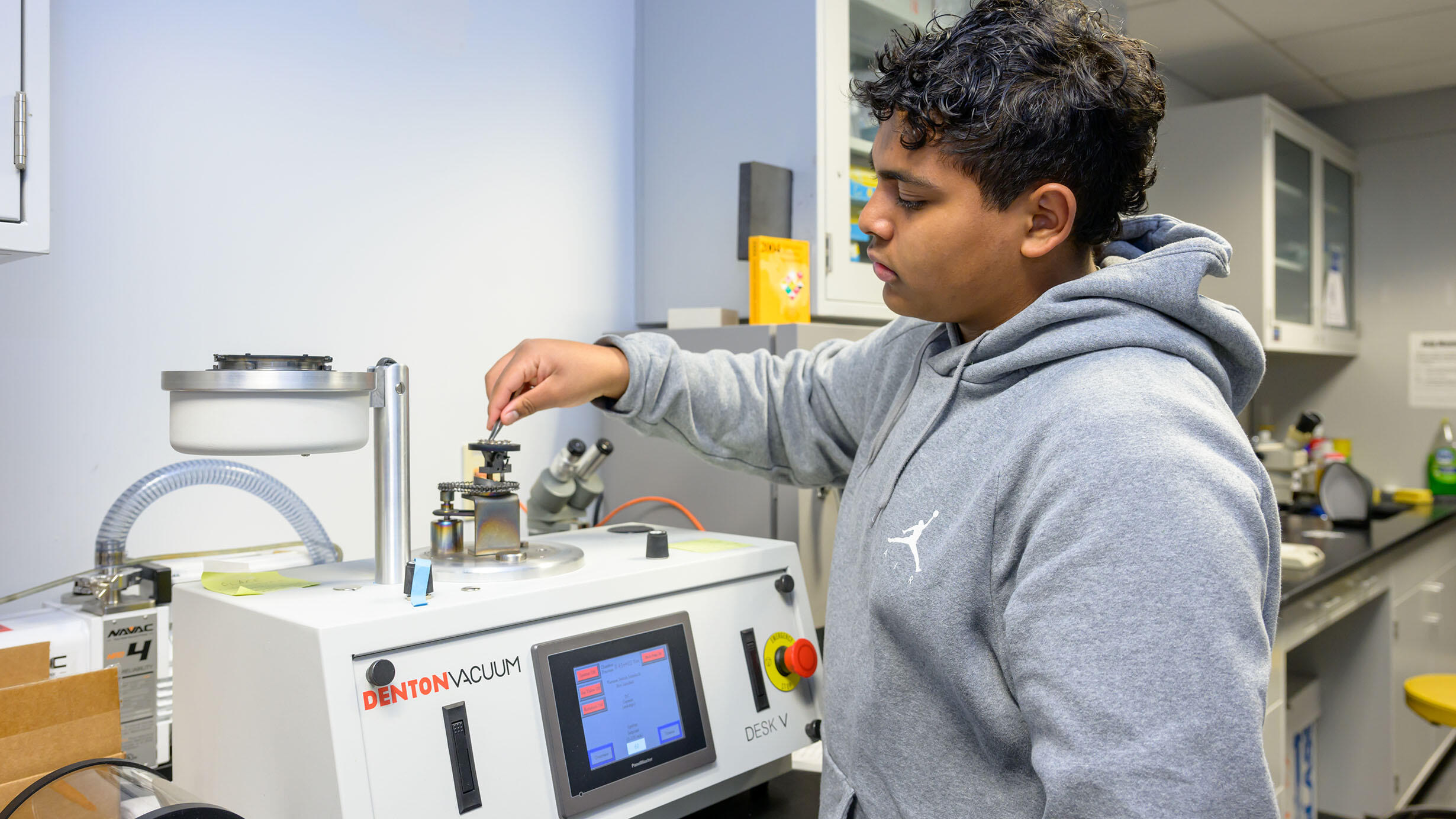 A student stands in front of a Denton Vacuum machine in a lab. 
