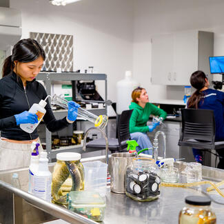 A student puts liquid from a squeeze bottle with a thin, curved opening into a beaker in a laboratory, with two more students seated in the background.