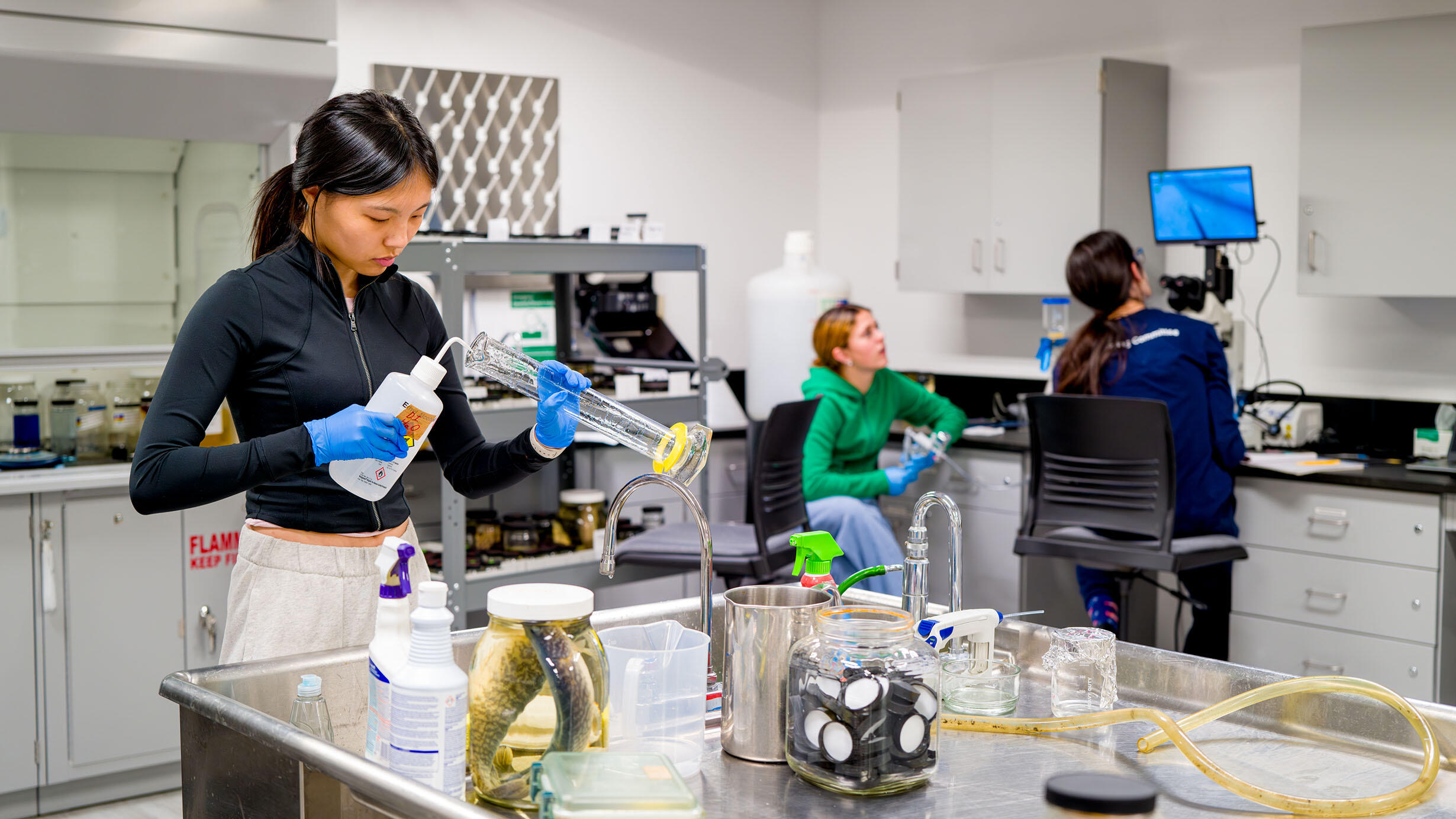 A student puts liquid from a squeeze bottle with a thin, curved opening into a beaker in a laboratory, with two more students seated in the background.
