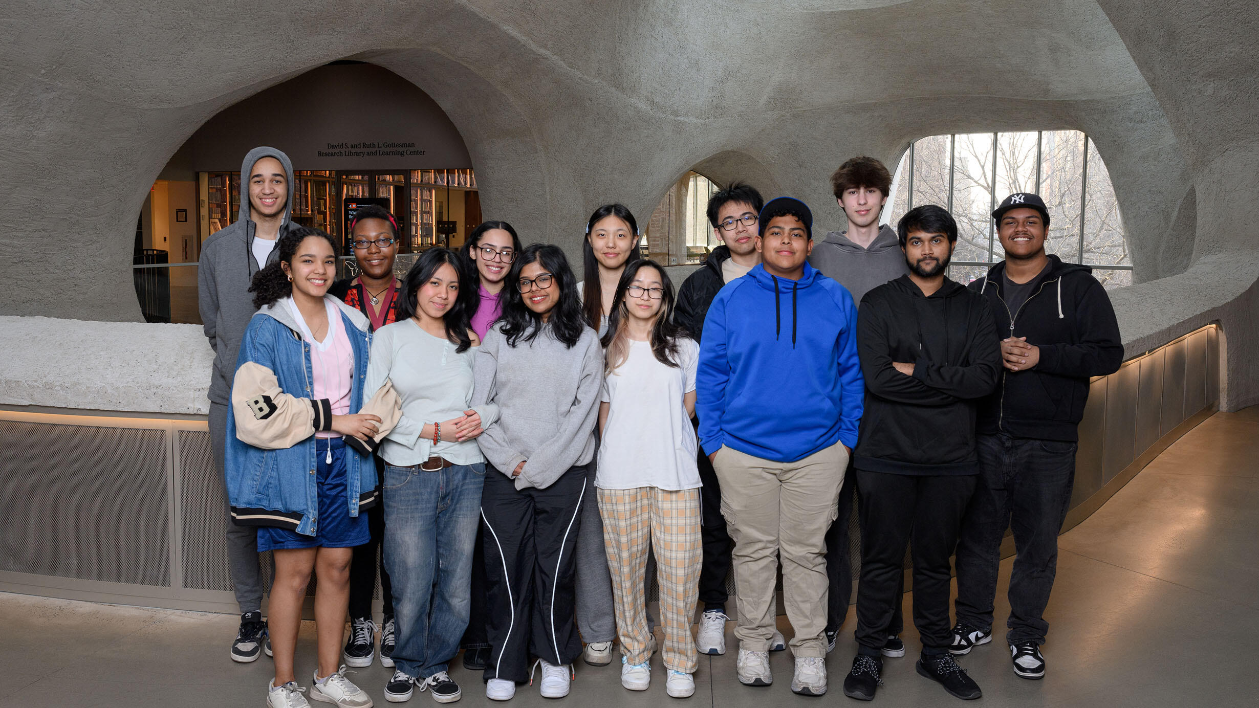Thirteen students stand in two rows in the Richard Gilder Center for Science, Education, and Innovation. 