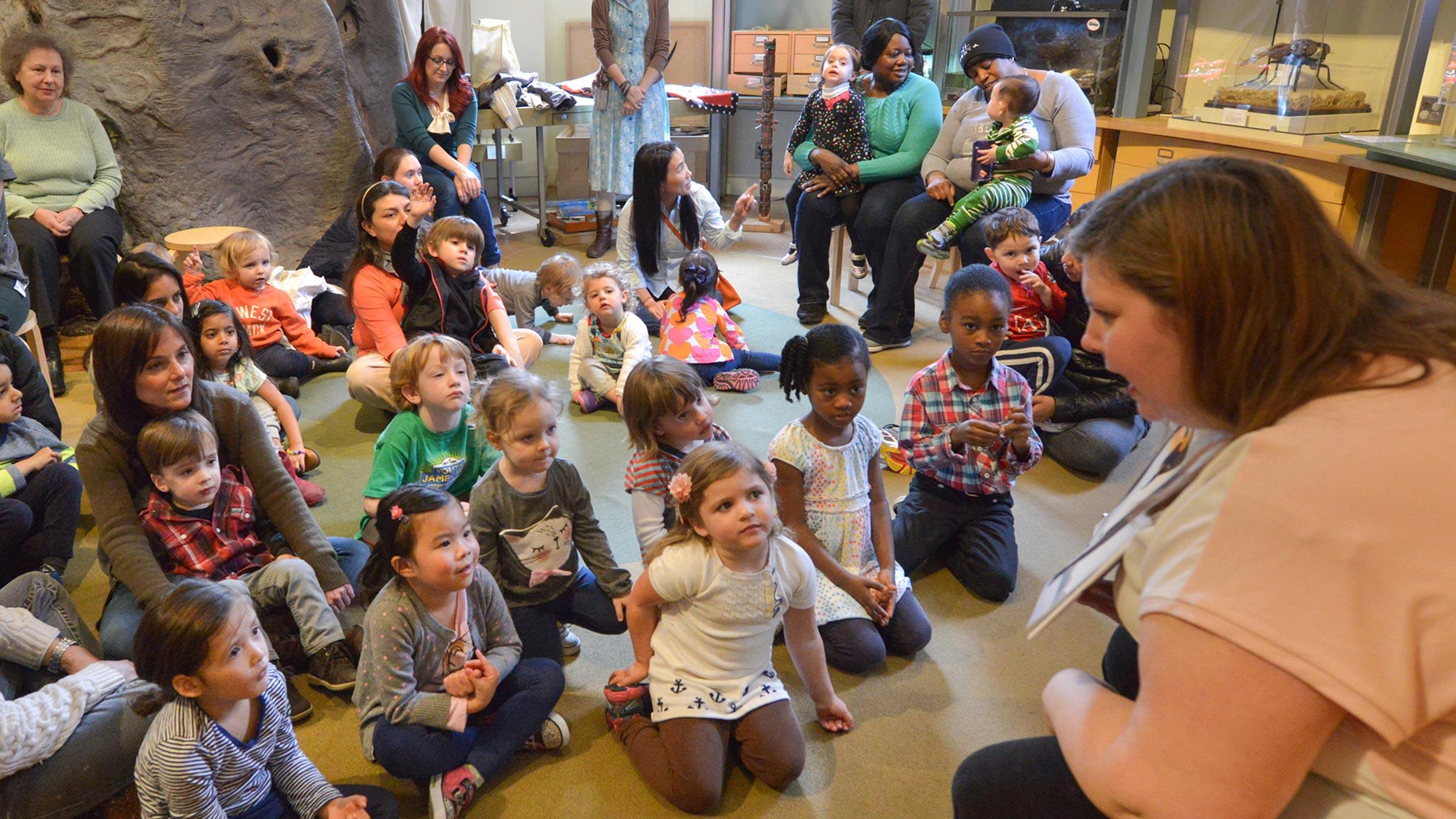 A seated Museum instructor leans over and speaks to a classroom of about twenty children and a dozen adults, most of whom are seated on the floor.