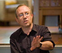 Caleb Scharf wearing a black shirt and glases and holding out his arm to the viewer as if explaining a thought