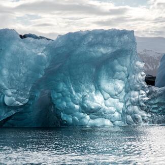 Large iceberg looming over water with snow-capped mountains in background and cloudy sky above.