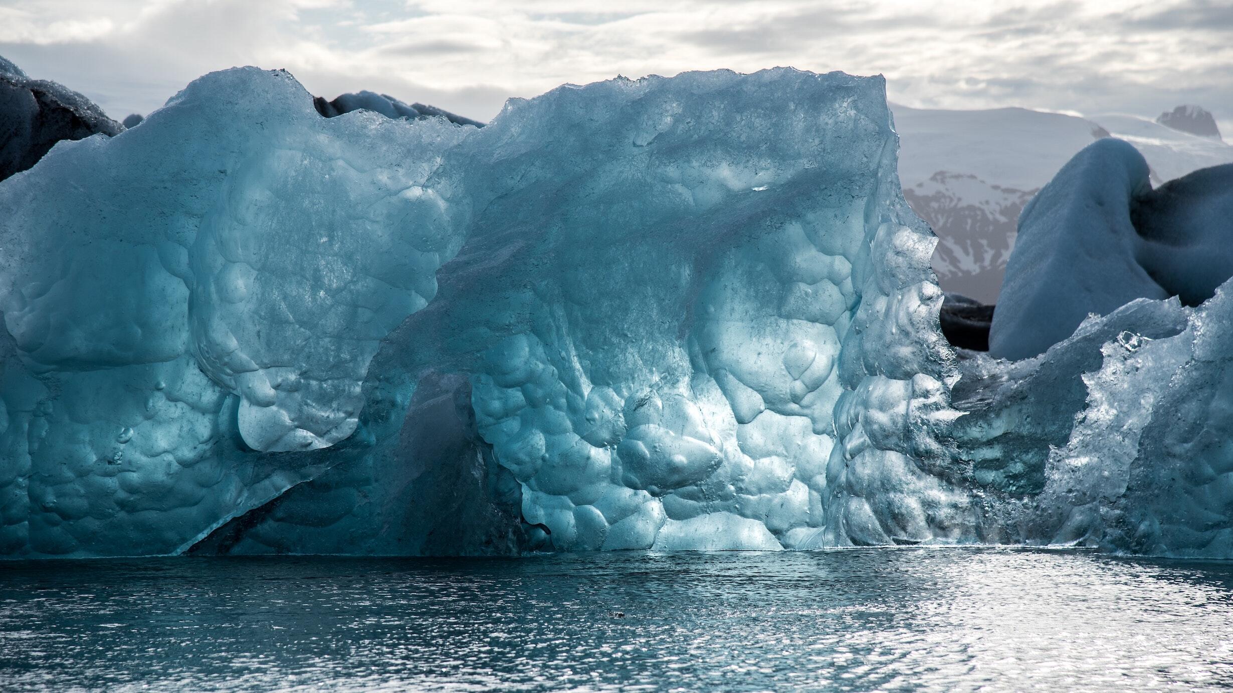 Large iceberg looming over water with snow-capped mountains in background and cloudy sky above.