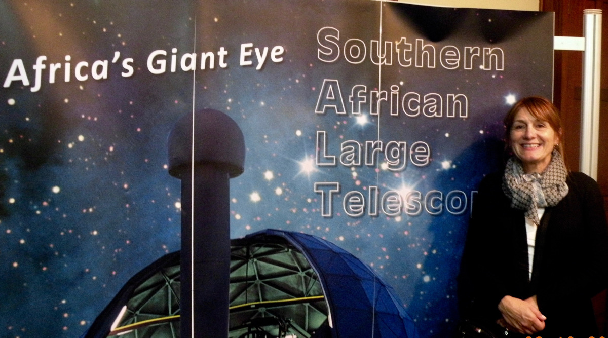 Mariel O'Brien standing in front of a large poster of a starlit sky that says "Africa's Giant Eye - Southern African Large Telescope"