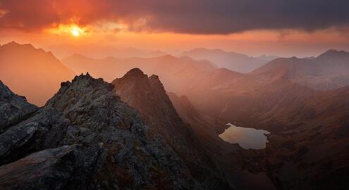 View of mountains in the foreground and stretching into the background, a lake in a valley below, and a multicolor sunset above.