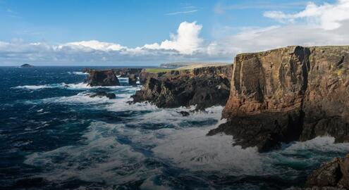 Seaside cliffs above rough water and a bright sky above, mostly clear with a large low cloud formation.