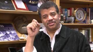 A man, Neil deGrasse Tyson, in an office with books and plaques on the shelves behind him.
