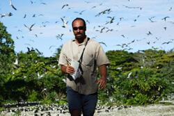 A man in the field outdoors facing the camera, with a flock of birds overhead and green vegetation in the background.