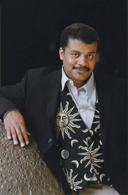 Neil deGrasse Tyson leaning on a large rock with a pitted surface.