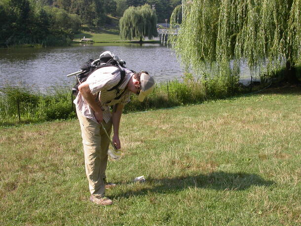 Dr. Noah Fierer, wearing a baseball cap and backpack, bends down while placing a specimen in a plastic bag in Central Park.