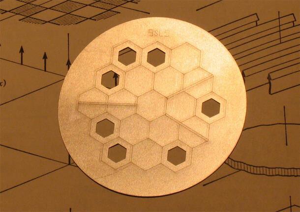 A disc whose surface is etched with the outlines of 19 small hexagons. Seven of those are perforated at irregular distances.