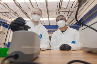 Two researchers wearing protective gear inside the abLab place a specimen in a vortexer.