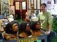 In a Museum collection room woman stands beside a rolling cart containing model busts of hominids with dark head hair and heavy brow ridges.