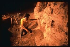 A man at an excavation site examining a stone wall that has small sections cut out.