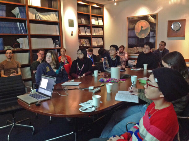 A group of people, seated around a large oval conference room table, watching a presentation.