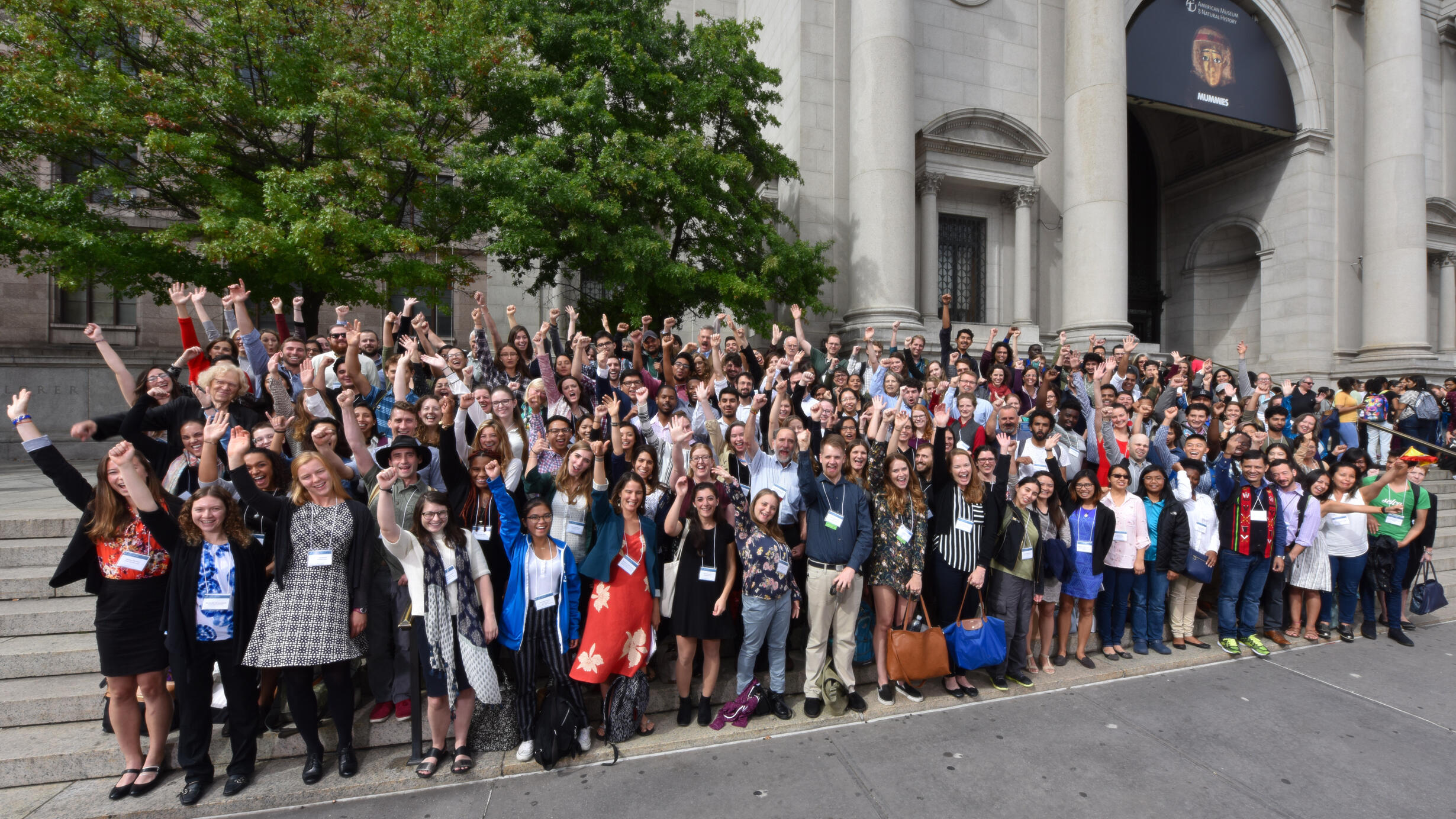 Over 150 Student Conference on Conservation Science participants pose on the Central Park West entrance steps, cheering with their right arms up.