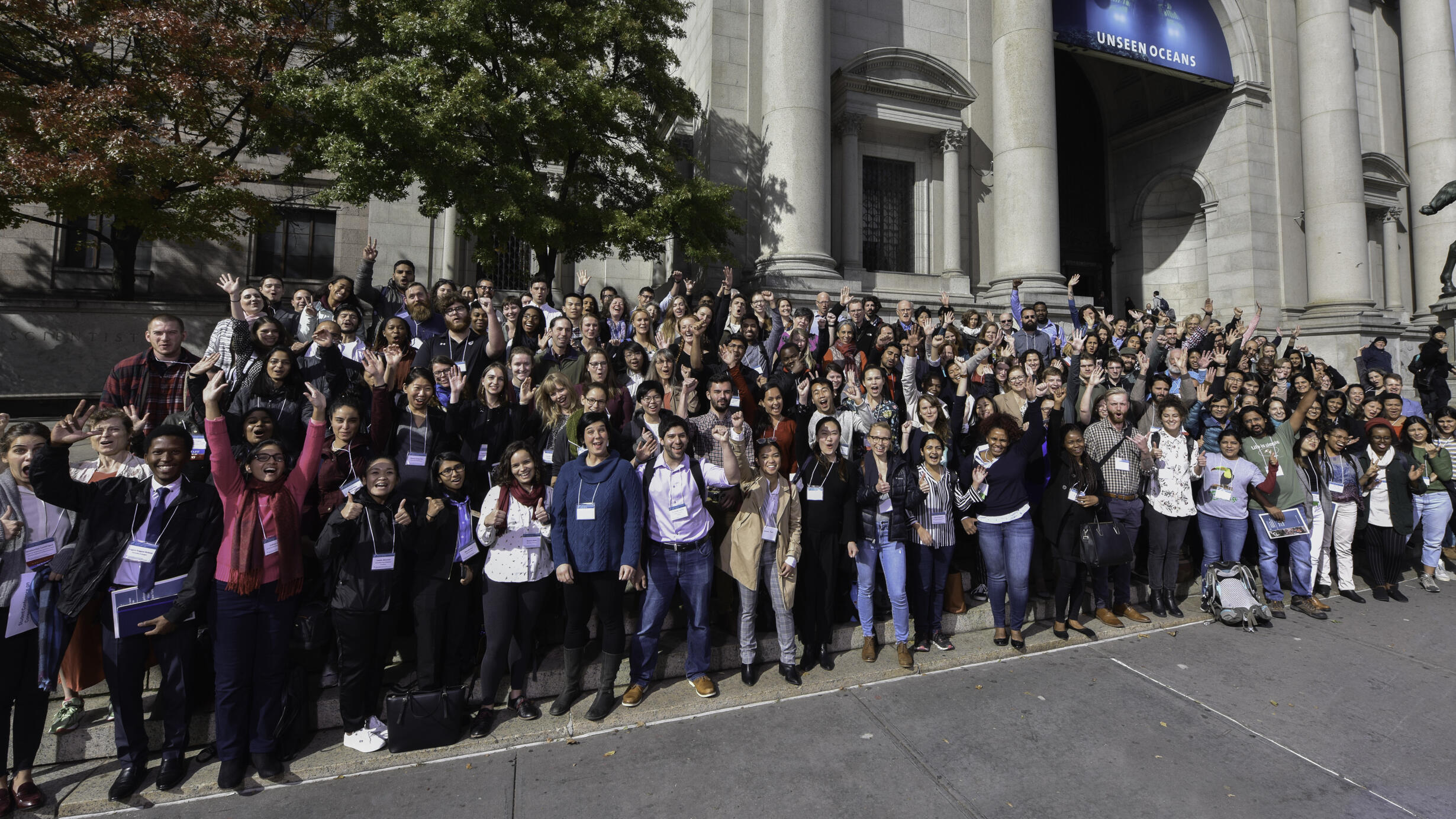 Over 150 Student Conference on Conservation Science participants pose on the Central Park West entrance steps, cheering with their right arms up.