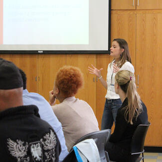 Person speaks in front of a conference room with four seated people visible.