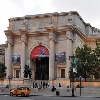 Central Park West entrance of the American Museum of Natural History in the daytime with a cab outside and people sitting on the steps.