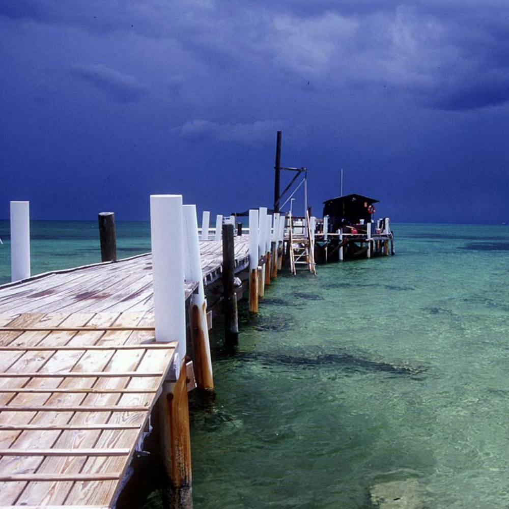 A pier with a building on the end stands in shallow clear water against a blue sky.