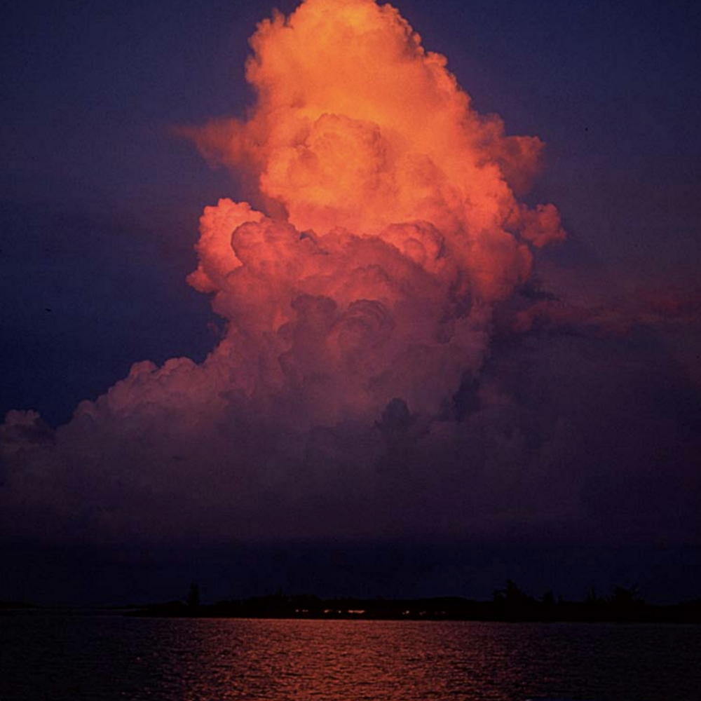 Billowing clouds above the ocean glow a dark red from the sunset.