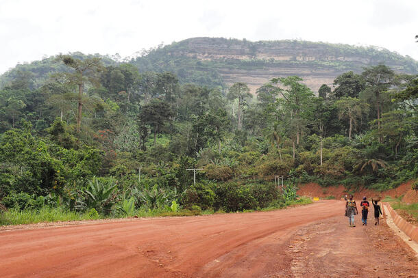Two people walk down a dirt road at the foot of a forested mountain.