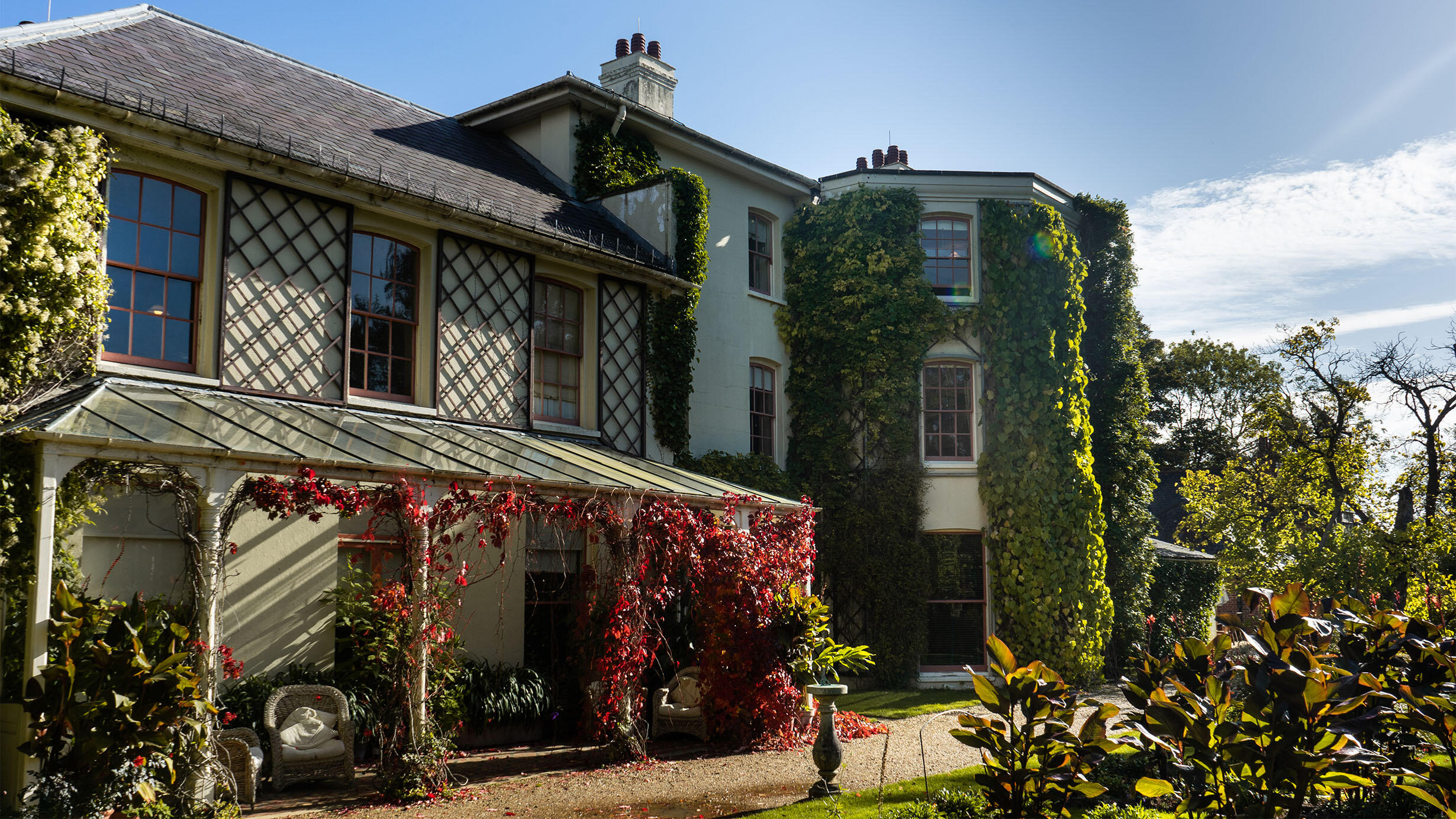 Large house with many windows, covered in greenery. A rounded corner and a glass roof patio which looks out on a garden are both covered in vines.