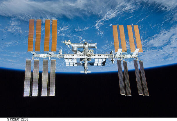 Modular International Space Station with long body and 16 solar arrays jutting out in orbit above Earth.