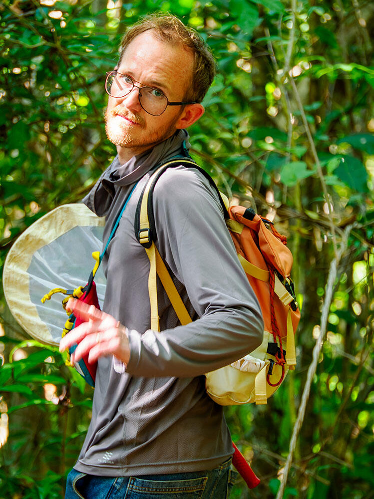 Corey Smith walks through a wooded area wearing a backpack and carrying collection equipment.
