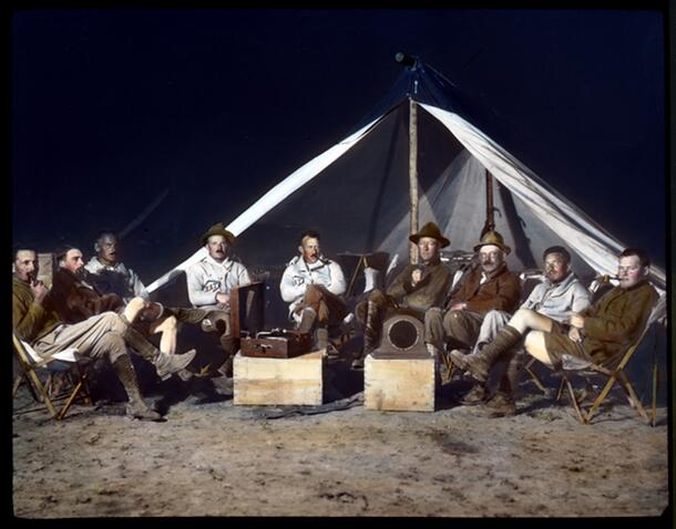 Period photo of nine men in field clothes, seated in semicircle on folding chairs outside a tent, on sandy soil, some with pipes, with a Victrola and a speaker perched on crates.